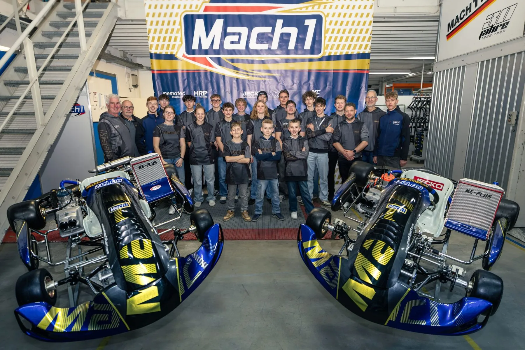 Das Mach1 Team in Mach1 Teamwear vor dem Mach1-Banner, links und rechts je ein Zelos Rennkart